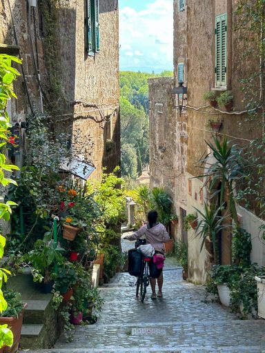 Ciclista sulla Via Francigena tra case in pietra e piante, nel centro storico di Capranica.