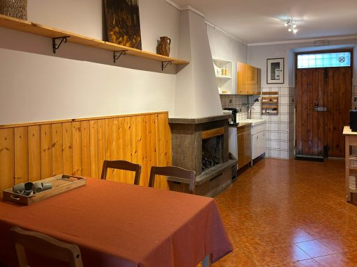 Vista dal tavolo da pranzo verso la porta d’ingresso e l’angolo cucina, con camino rustico.