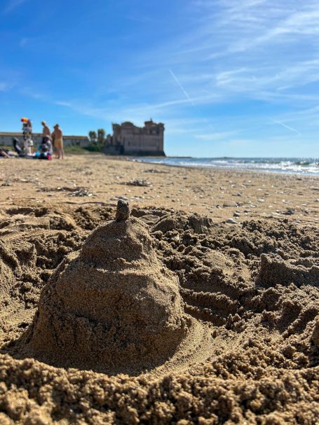 Castello di sabbia sulla spiaggia di Santa Severa, con il castello vero sullo sfondo e il mare sotto il cielo azzurro.