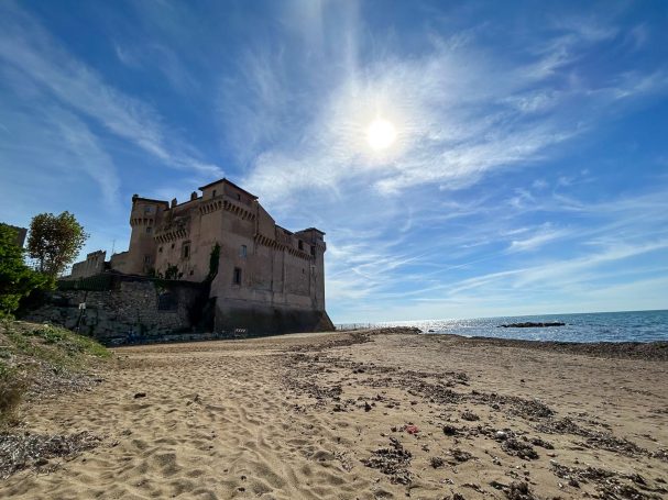Castello di Santa Severa visto dalla spiaggia, con il mare sullo sfondo e il cielo azzurro di una giornata mediterranea.