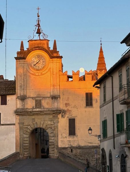 Porta dell’Orologio a Capranica al tramonto, con il centro storico sullo sfondo e la luna che sorge tra i tetti medievali.